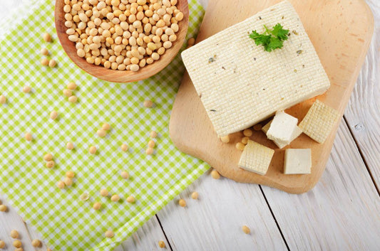 duzy kawalek tofu polozony na drewnianej desce obok miseczki z ziarnami soi, na drewnianym stole i kuchennej sciereczce