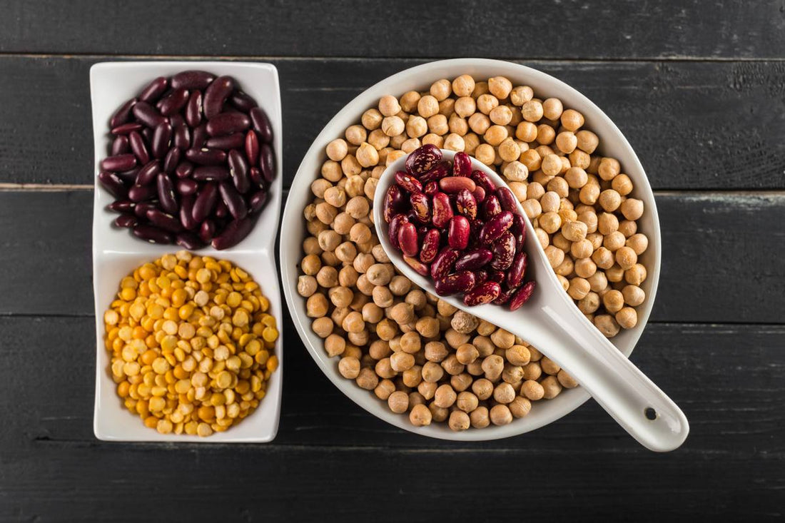 duża biała miska na drewnianym stole, wypełniona ziarnami soi, na misce położona biała duża łyżka wypełniona czerwoną fasolą, obok dwie miseczki wypełnione czarną fasolą i soczewicą