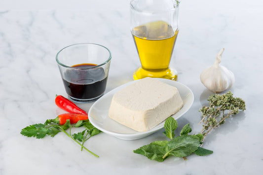 kostka tofu na białym talerzyku, położona na marmurowych blacie, otoczona przez papryczki chilli, oliwę, sos sojowy i zioła