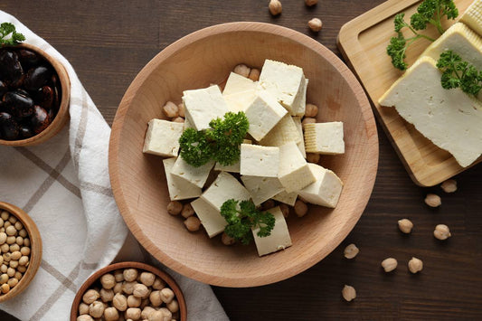 kawałki tofu ułożone w glinianej misce, koperkiem, postawione na drewnianym stole, w towarzystwie ziaren soi i kuchennej ścierki